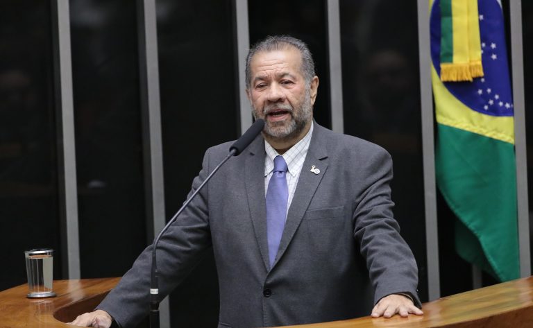 Um homem branco com cabelo e barba grisalhos fala ao microfone. Ele usa terno cinza e ao fundo, na direita, tem uma bandeira do Brasil
