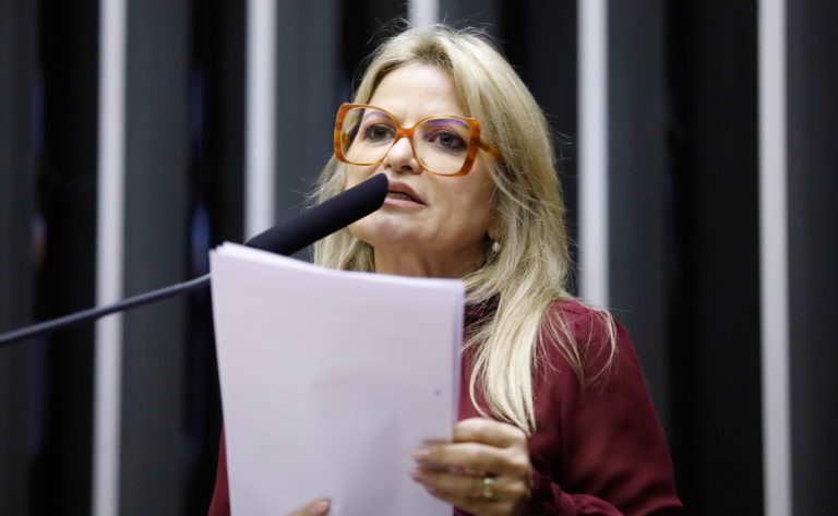 Deputada Flávia Morais fala ao microfone. Ela é loira, tem o cabelo comprido, usa óculos e uma blusa vermelha e segura um papel