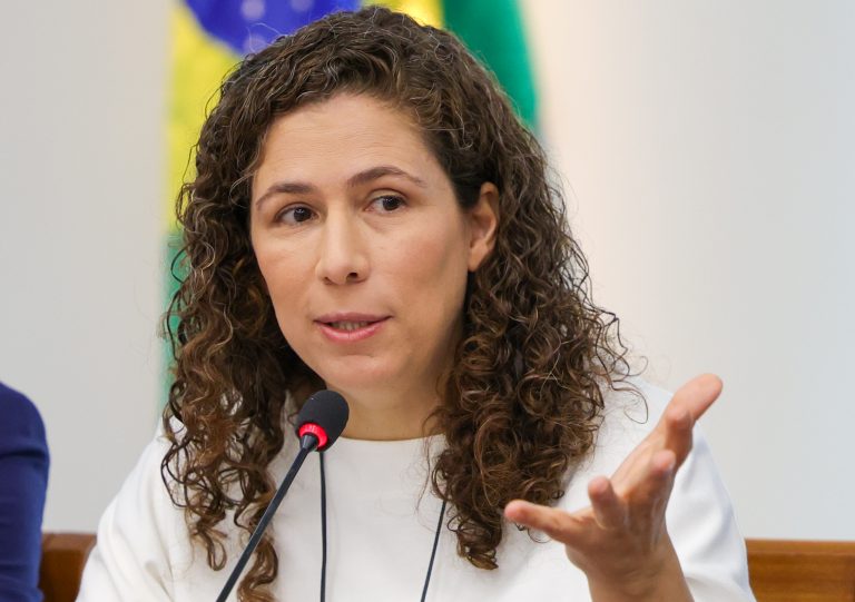 A ministra Esther Dweck fala ao microfone. Ela é uma mulher branca, tem cabelo castanho encaracolado passado dos ombros, e veste uma camisa branca