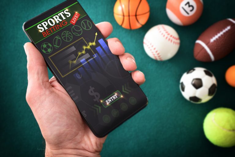 Uma pessoa segura um celular que tem na tela um aplicativo de apostas. No fundo há bolas de várias modalidades esportivas
