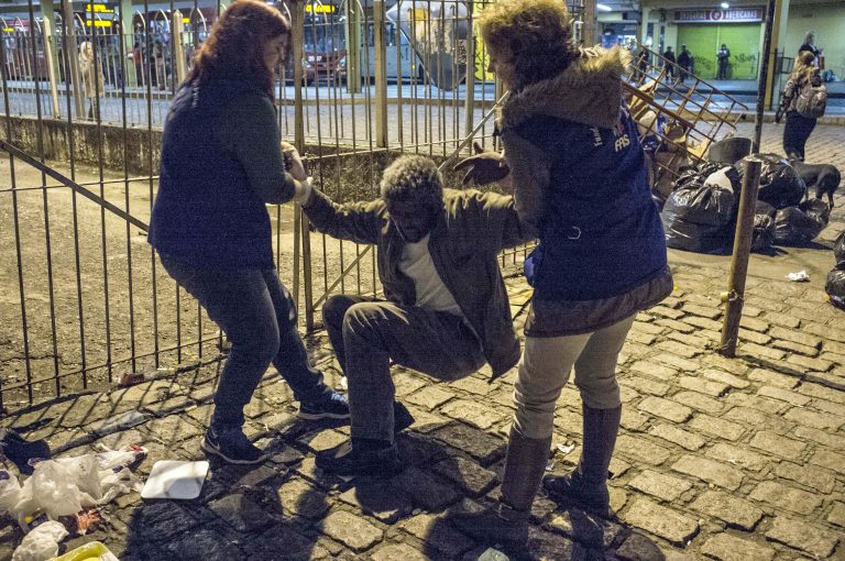 Duas mulheres tentam levantar um morador de rua