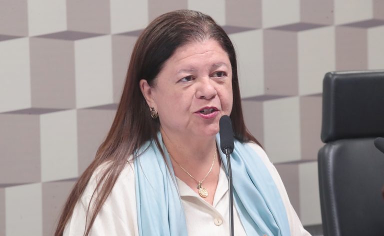 Deputada Laura Carneiro fala ao microfone. Ela é branca, tem o cabelo comprido e escuro, veste uma blusa branca com uma echarpe azul clara