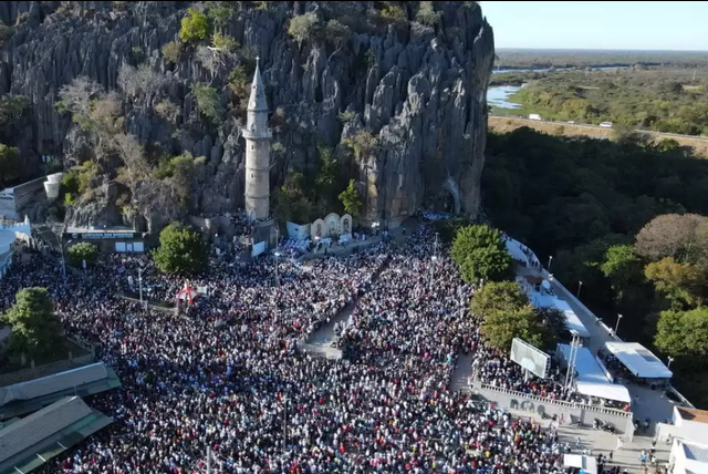 Foto aérea de uma procissão, Ao fundo, há um morro e a torre de uma igreja. O dia etá ensolarado
