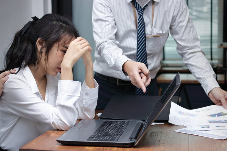Mulher jovem está sendo constrangida no trabalho. Um homem aponta o dedo para a tela do laptop dela e ela está triste com a cabeça baixa e as mãos no rosto