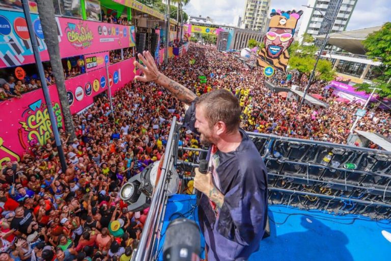 Um cantor está em cima de um trio elétrio. Abaixo uma multidão segue o trio. Ele canta de frente para alguns camarotes. O dia está ensolarado