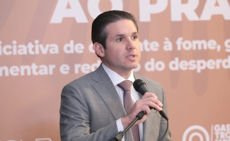 Hugo Motta fala ao microfone. Ele é um homem branco, tem o cabelo escuro, veste um terno azul-marinho com camisa branca e gravata vermelha