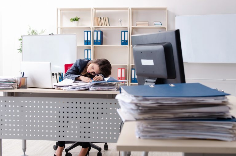 Mulher dorme em com a cabeça em cima da mesa de trabalho, que está cheia de papéis