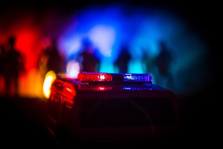 Carro de polícia com a luz ligada. Ao fundo há vultos de pessoas