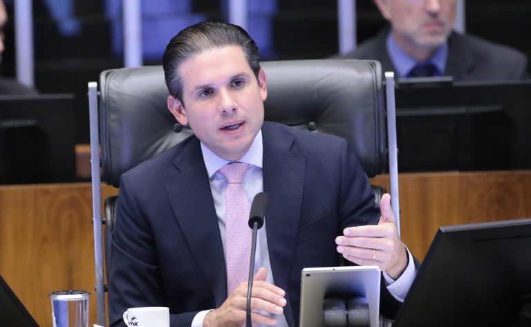 Hugo Motta fala ao microfone. Ele é um homem branco, tem o cabelo escuro, veste um terno azul-marinho com camisa branca e gravata vermelha