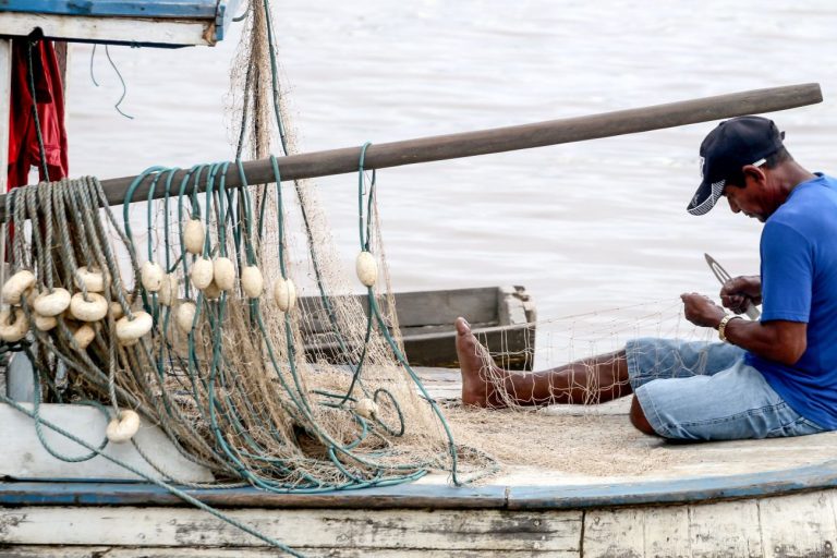 Pescador sentado num barco conserta a rede de pesca. Ele veste uma camisa azul e um boné preto