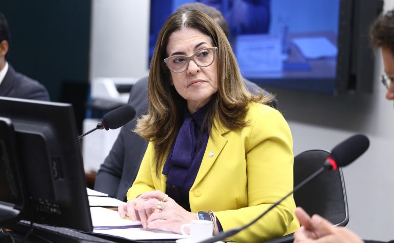 Deputada Rogéria Santos fala ao microfone. Ela é uma mulher clara, com cabelo castanho médio, usa óculos e blazer amarelo e uma camisa azul-marinho 