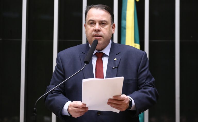 Deputado Julio Cesar Ribeiro fala ao microfone. Ele é um homem claro, com cabelo escuro, usa um terno azul-marinho, camisa branca e gravata vermelha. Ele segura um papel. No fundo, à direita, há uma bandeira do Brasil