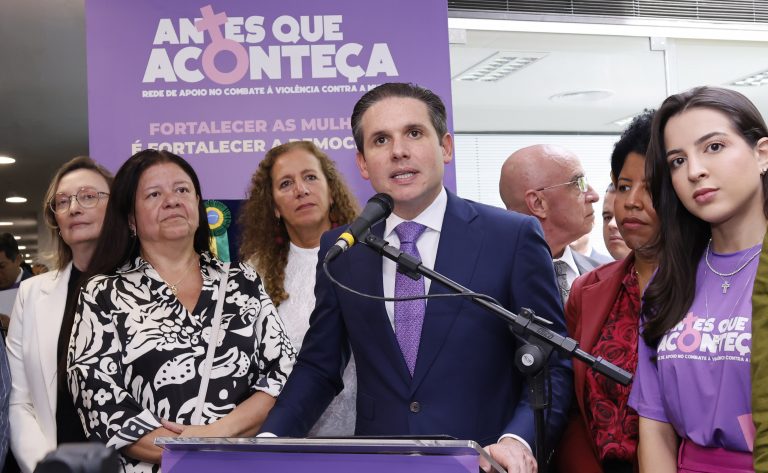 Deputados reunidos na frente de um painel lilás