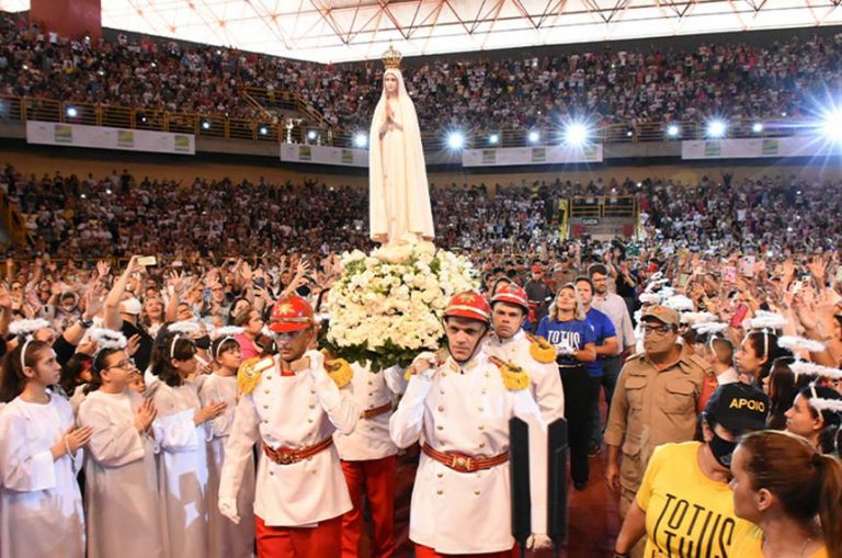Imagem de uma procissão religiosa em ginásio lotado, com uma imagem de Nossa Senhora sobre um andor decorado com flores brancas.Homens uniformizados carregam o andor enquanto fiéis, crianças vestidas de branco e equipes de apoio acompanham o cortejo.