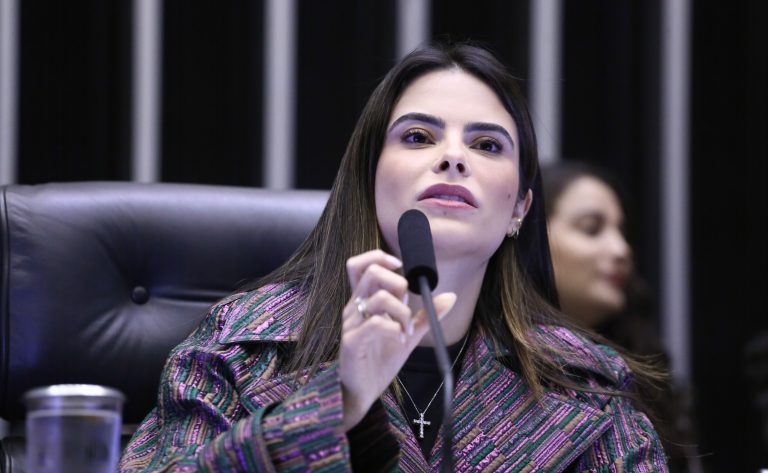 Deputada Yandra Moura fala ao microfone. Ela é uma mulher branca, tem cabelo longo, liso e castanho, e veste um blazer escuro