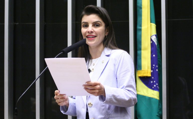 Deputada Marussa Boldrin fala ao microfone. Ela é branca, tem cabelo comprido, liso e escuro; veste um blazer claro e segura um papel. Ao fundo, à direita, há uma bandeira do Brasil num mastro