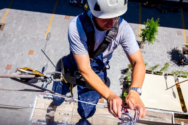 Fotografia vista de cima mostra um trabalhador em altura, usando capacete branco, uniforme azul e cinto de segurança preso por cordas e mosquetões. Ele trabalha na parte externa de um edifício, segurando uma estrutura metálica junto à fachada. Ao fundo, vários metros abaixo, aparecem calçada, árvores, postes e veículos.
