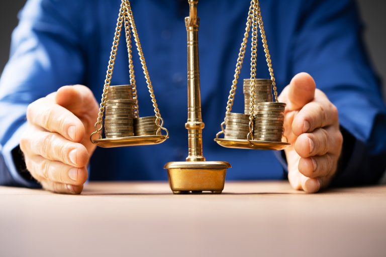 Pessoa com as palmas das mãos em cima de uma superfície, voltadas para dentro, aonde há uma balança com moedas em seus pratos 