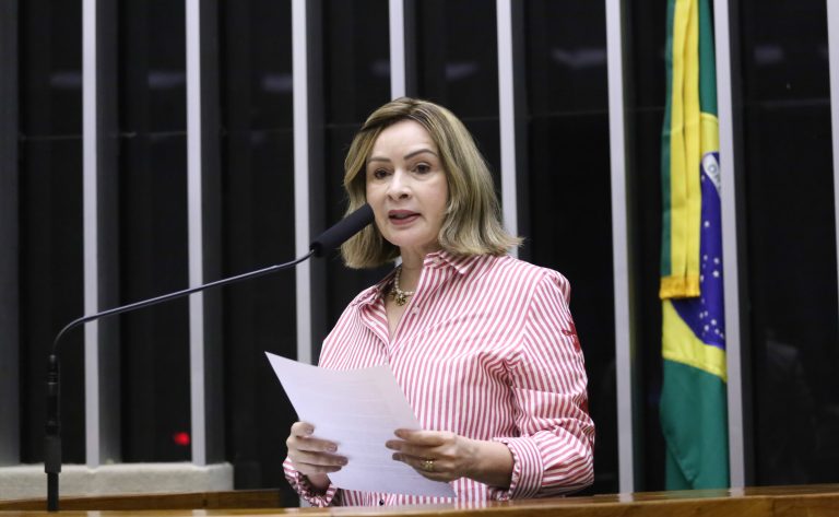 Deputada Socorro Neri fala ao microfone. Ela é uma mulher branca, tem o cabelo liso, loiro, na altura dos ombros, veste uma camisa listrada de vermelho e branco e segura um papel
