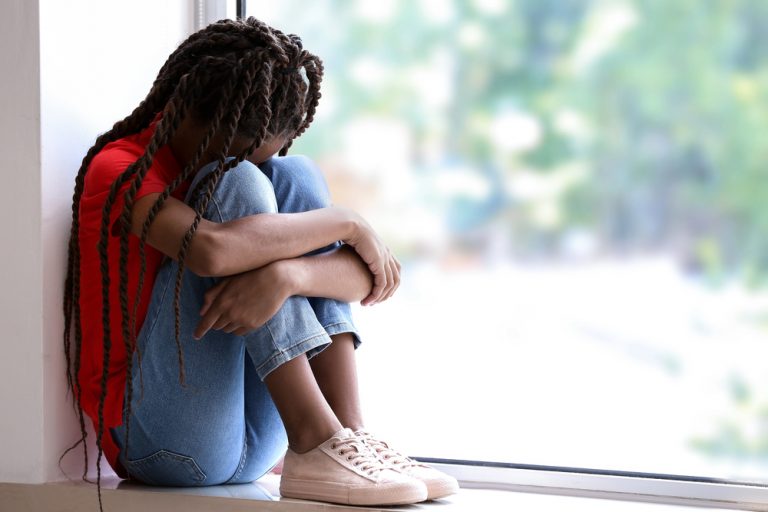 Menina negra, racismo, preconceito racial, discriminação – Menina negra sentada no chão, em forma de concha, com as pernas dobradas e apertadas contra o tronco e o rosto escondido sobre os joelhos