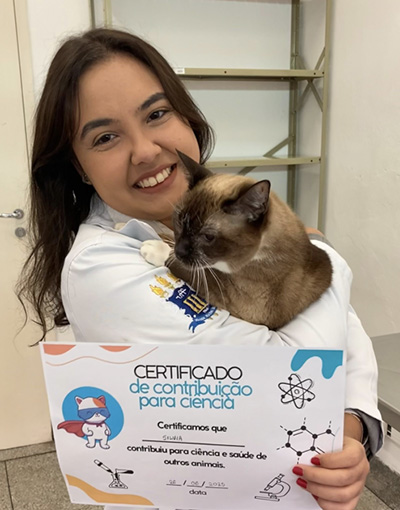 Pesquisadora, usando jaleco branco, segura no colo um gato siamês enquanto apresenta um certificado de contribuição para a ciência. O documento agradece a participação do animal na pesquisa e destaca sua contribuição para a ciência e a saúde de outros animais.