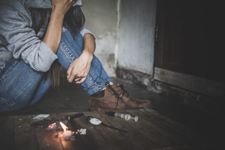 Foto de uma mulher com a cabeça abaixada e sentada no chão. Na frente, no chão há drogas, um isqueiro e uma seringa