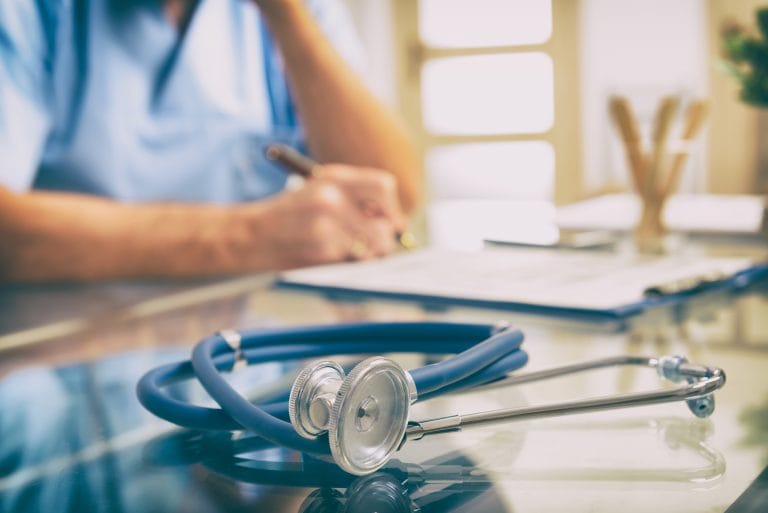 Foto cortada e desfocada de um médico com um jaleco azul claro, em cima da mesa há um estetoscópio