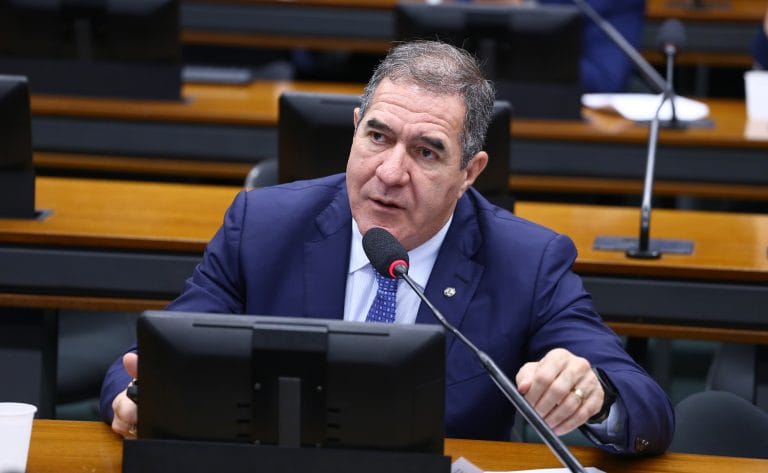 Deputado Luiz Gastão fala ao microfone. Ele é um homem claro, usa um terno azul marinho e camisa branca