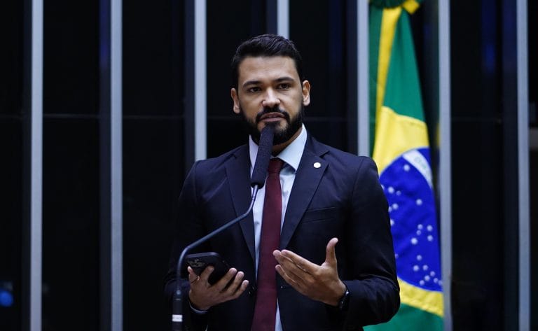 Deputado Samuel Viana fala ao microfone. Ele é um homem claro com cabelo e barba pretos, usa um terno azul escuro e ao fundo no lado direito há uma bandeira do Brasil