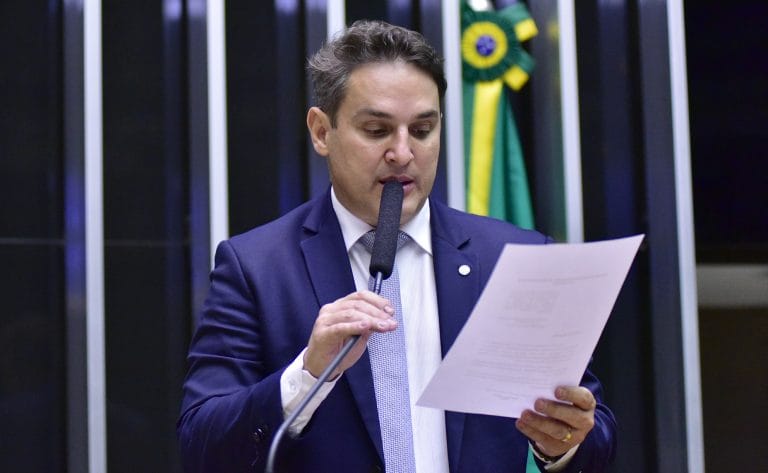 Deputado Zucco fala ao microfone. Ele é um homem claro, com cabelo curto e escuro. Veste um terno azul-marinho e segura um papel