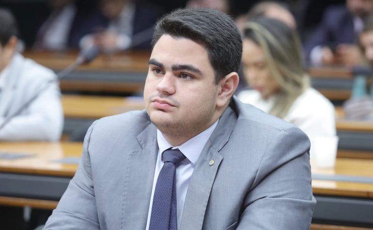 Deputado Ícaro de Valmir fala ao microfone. Ele é branco, tem cabelo escuro e veste um terno cinza-claro