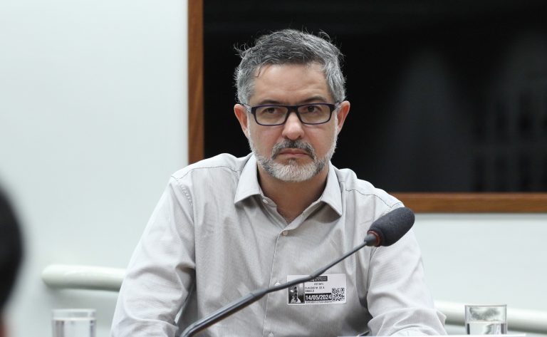 Um homem claro com cabelos grisalhos olha pra frente. Ele usa óculo e uma camisa cinza claro.