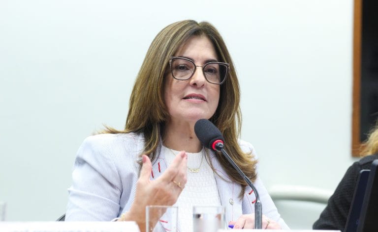 Deputada Rogéria Santos fala ao microfone. Ela é clara, tem cabelo liso, comprido e castanho claro. Ela usa um blazer claro e óculos