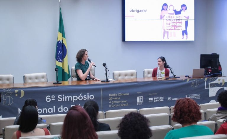 Duas mulheres estão sentadas à mesa, enquanto uma delas fala ao microfone