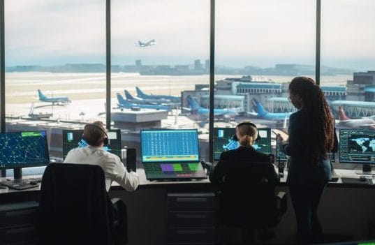 Diversas equipes de controle de tráfego aéreo trabalhando em uma Torre de Aeroporto Moderno. A Sala de Escritório está cheia de monitores de computador de mesa com telas de navegação, dados de partida e chegada de aviões para controladores.