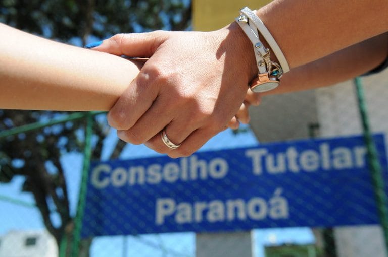 Uma mulher e uma criança de mãos dadas. Ao fundo, uma placa escrito: Conselho Tutelar Paranoá