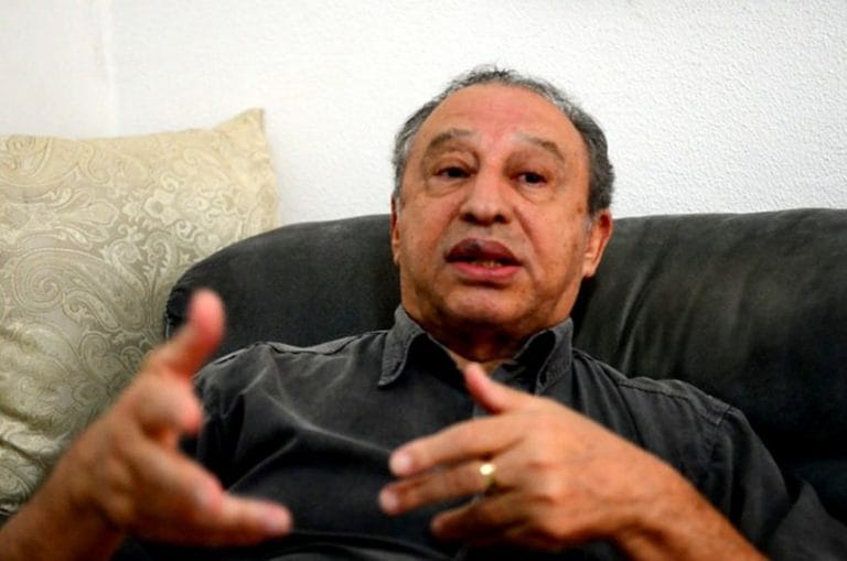 Um homem está recostado no sofá e fala olhando para a frente