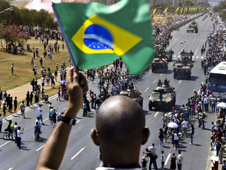 Pessoas desfilam na rua, em primeiro plano um homem segura uma bandeira do Brasil