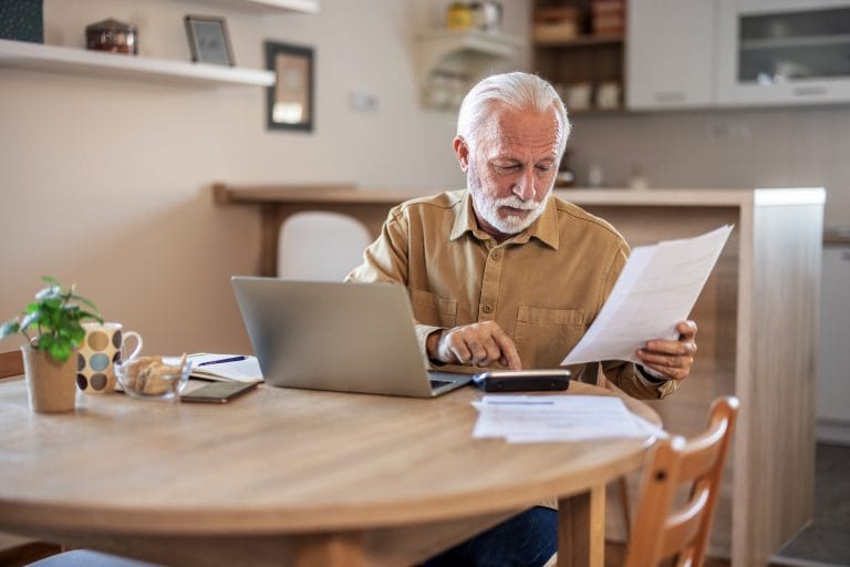 Um idoso faz cálculos com um papel na mão e uma calculadora. Em cima da mesa ha um laptop. Ele tem cabelo, barba e bigode branco, e veste uma camisa cáqui