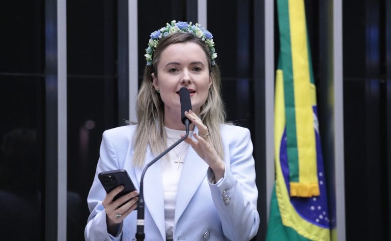 Deputada Julia Zanatta fala ao microfone. Ela é branca, loira, usa um blazer branco, uma tiara de flores na cabeça e segura um celular
