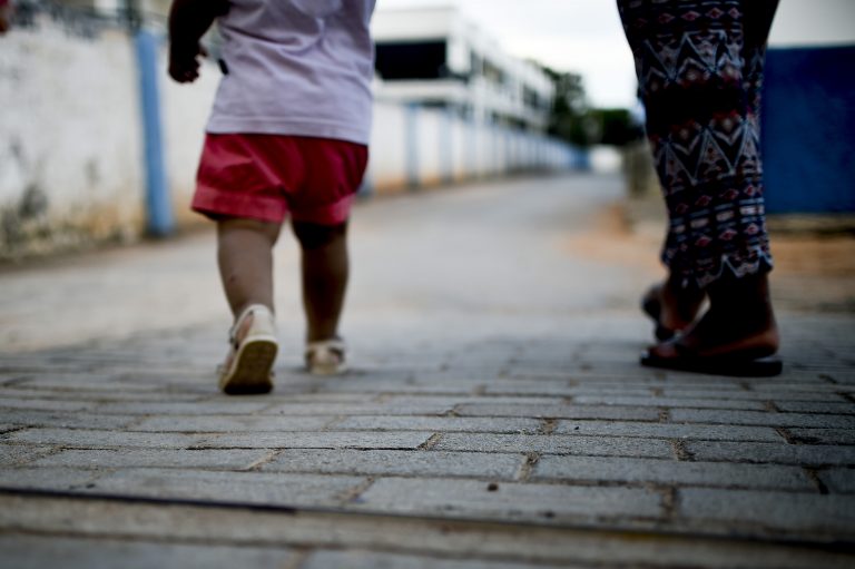 Mãe e filho bebê caminham num rua vazia. Eles estão de costas e a foto está cortada na altura da cabeça da criança