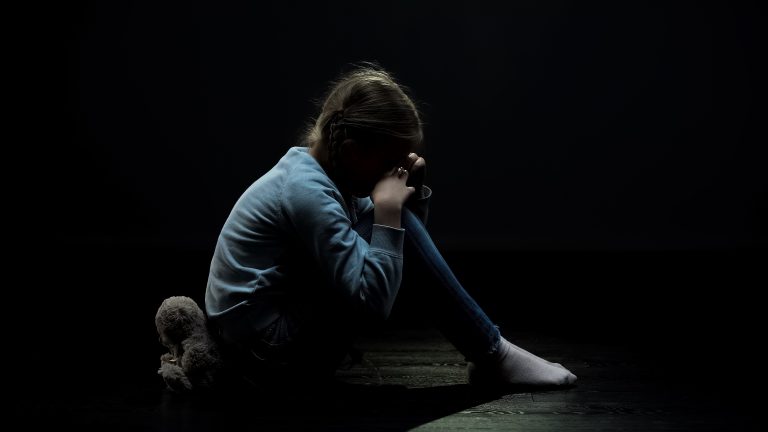 Uma menina está sentada no chão com a cabeça apoiada nos joelhos, está escuro e não é possível ver o rosto da criança