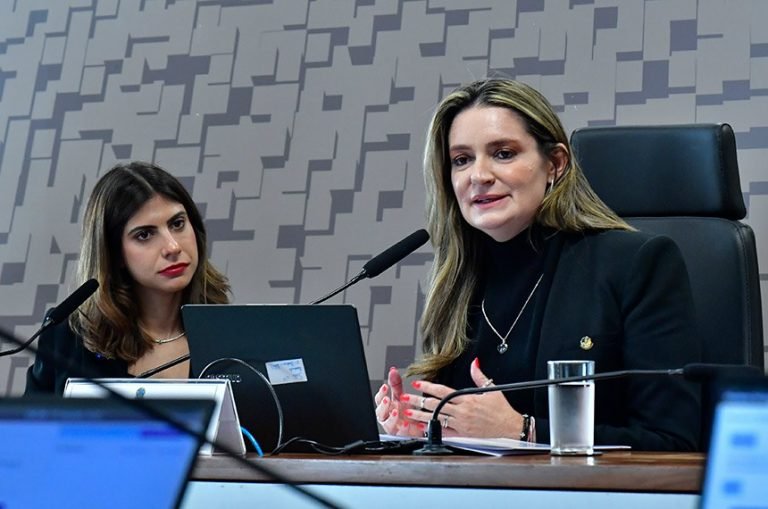 Duas mulheres estão sentadas à mesa, enquanto uma delas fala ao microfone