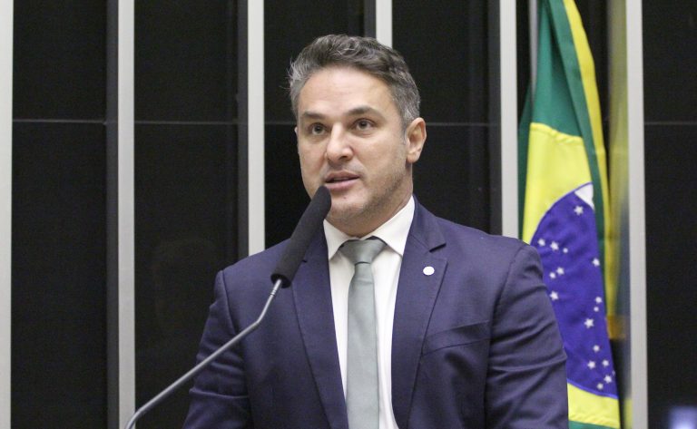 Deputado Zucco fala ao microfone. Ele é um homem claro, com cabelo curto e escuro. Veste um terno azul-marinho e segura um papel