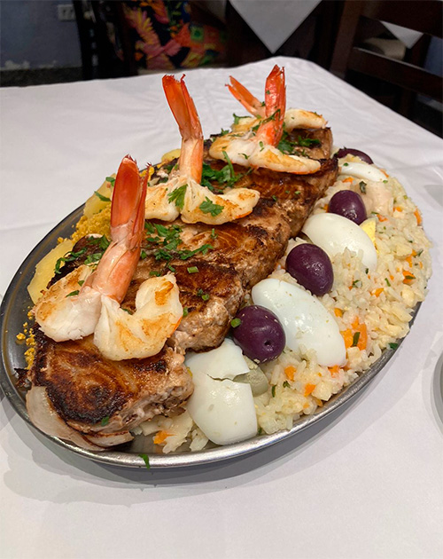 foto montagem de alimentos: travessa de alumínio contendo bife grande sobreposto de camarões, acompanhado de arroz, ovos e azeitona e outro prato de louça branca contendo lagosta frita, acompanhado de batatas e salada.
