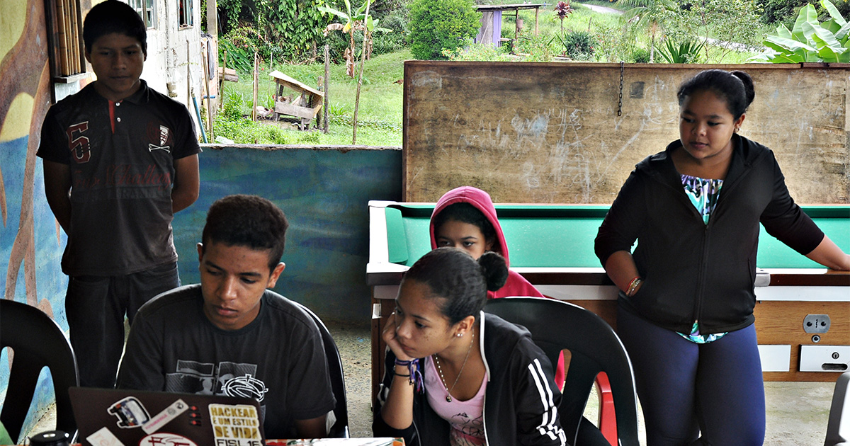 Cinco jovens se reúnem em volta de um notebook, na varanda de uma casa rural. Uma das meninas se apoia em uma mesa de sinuca. No quintal, ao fundo, há bananeiras e diversas outras espécies de plantas.