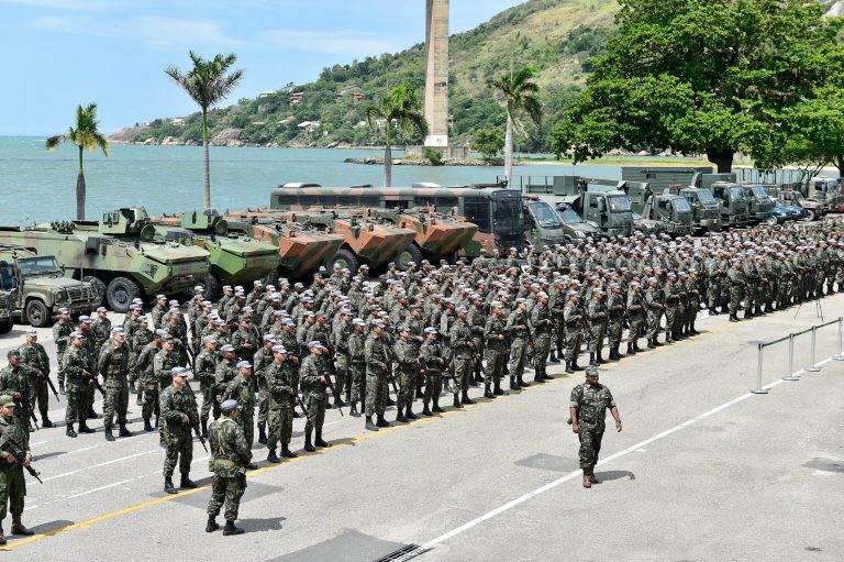 Num dia de sol, vários soldados do Exército estão em pé, em filas, segurando armas. Atrás deles há tanques de guerra, e no fundo se vê o mar