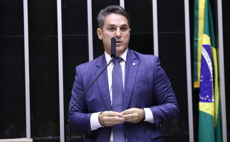 Deputado Zucco fala ao microfone. Ele é um homem claro, com cabelo curto e escuro. Veste um terno azul-marinho